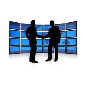 An image of two businessmen shaking hands with a flat world map on multiple screens behind them.