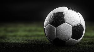 A close-up of a textured black and white soccer ball on a green field with a blurred dark background.

