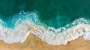 Aerial view of turquoise waves crashing on a sandy beach, creating a serene coastal scene.

